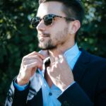 Fashionable man adjusting his suit and sunglasses outdoors on a sunny day.